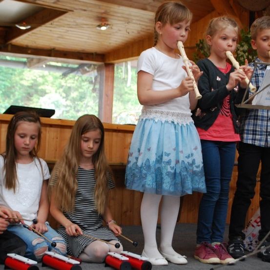 Mehrere junge Kinder stehen und sitzen in einem Raum und spielen Musikinstrumente. Ein Mädchen in einem blauen Rock bläst eine Flöte, während andere Xylophon spielen. Hinter ihnen spielt ein Mädchen die Flöte. Vor den Kindern steht ein Notenständer.