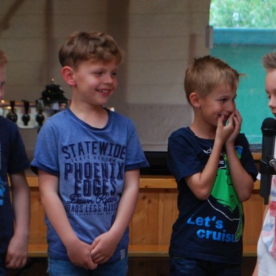 Vier Jungen stehen in der Nähe eines Mikrofons, einer spricht. Sie tragen lässige Hemden mit Designs. Hinter ihnen ist eine Holztheke mit verschiedenen Gegenständen.