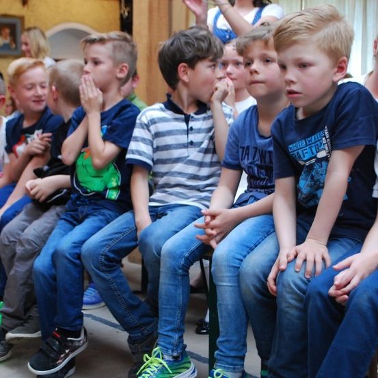 Eine Gruppe junger Jungen sitzt zusammen, trägt Freizeitkleidung, einige halten ihre Hände vor den Mund, möglicherweise in einem Klassenzimmer oder ähnlichem Umfeld.