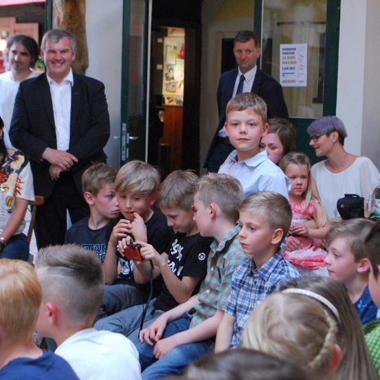 Mehrere Erwachsene und Kinder sind in einem Raum versammelt, wobei zwei Männer in der Nähe des Eingangs stehen. Ein Kind hält eine Kamera und hält den Moment fest. Der Raum hat eine Tür und Fenster.