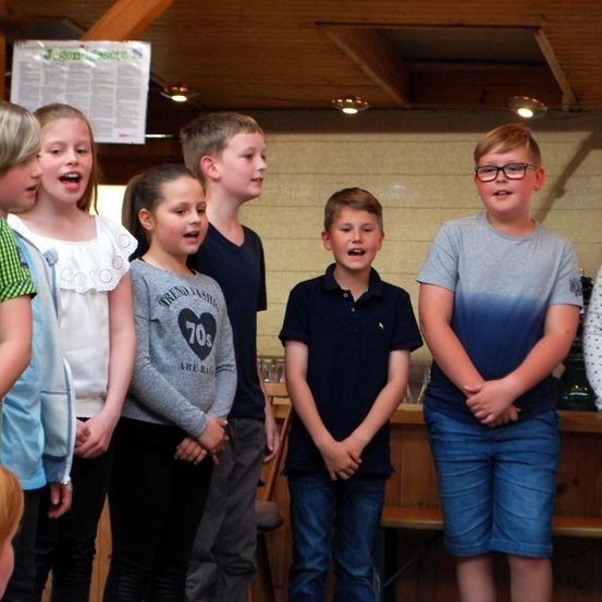 Eine Gruppe von Kindern steht in einem Raum und singt. Sie tragen Freizeitkleidung. Eine Wand ist hinter ihnen mit einem Plakat. Ein Holztresen ist im Vordergrund.