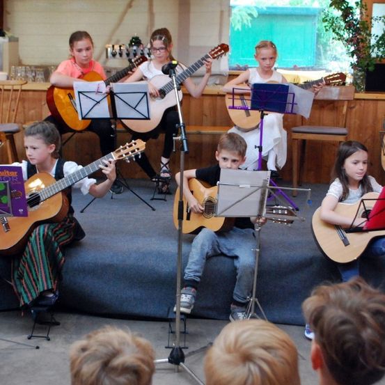 Mehrere Kinder spielen Gitarren auf einer Bühne vor einem Publikum. Einige haben Notenblätter, während andere nach unten schauen. Hinter ihnen steht eine Holztheke mit einer Pflanze.