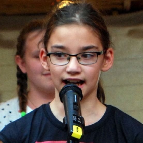 Ein junges Mädchen mit Brille singt in ein Mikrofon, mit einem weiteren Mädchen in einem gepunkteten Shirt im Hintergrund.