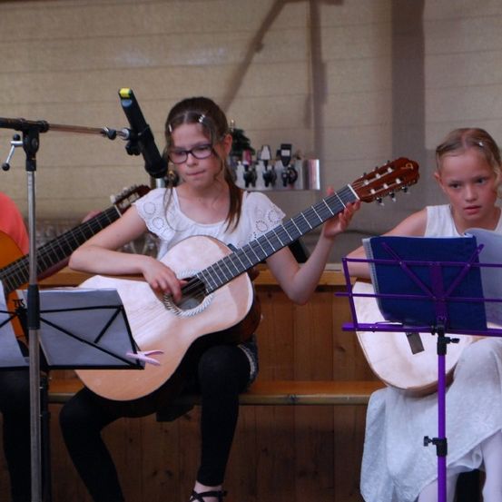 Drei Mädchen spielen Gitarre, wobei ein Mädchen in der Mitte eine Gitarre mit Mikrofon hält. Die anderen beiden haben Gitarren und Notenständer. Sie spielen wahrscheinlich auf der Bühne.