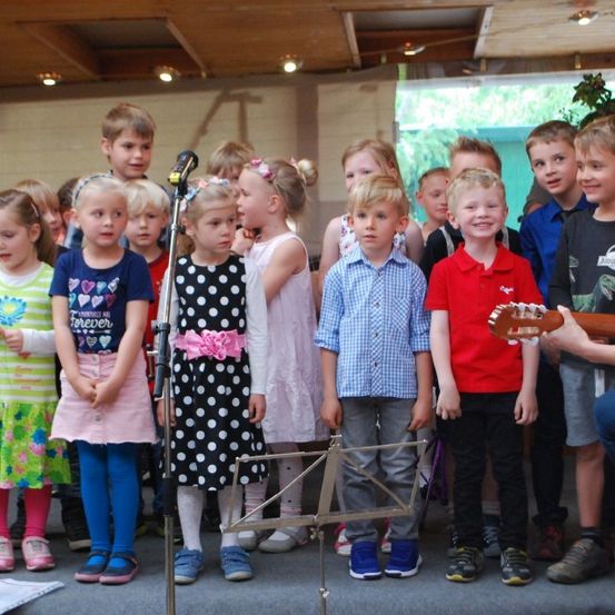 Eine Gruppe von Kindern steht vor einem Mikrofon mit einem Notenständer, ein Kind spielt Gitarre. Die Kinder tragen verschiedene Kleidungsstücke, darunter Kleider, Hemden und Hosen.