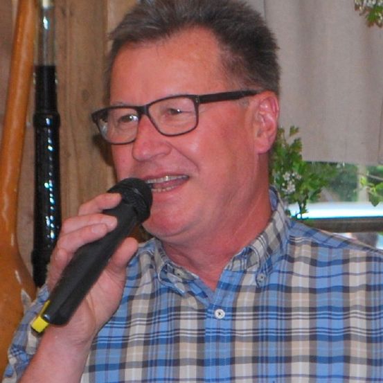 Ein Mann mit Brille hält ein Mikrofon und lächelt. Er trägt ein blaues kariertes Hemd. Hinter ihm ist eine Holzwand und ein Vorhang zu sehen.