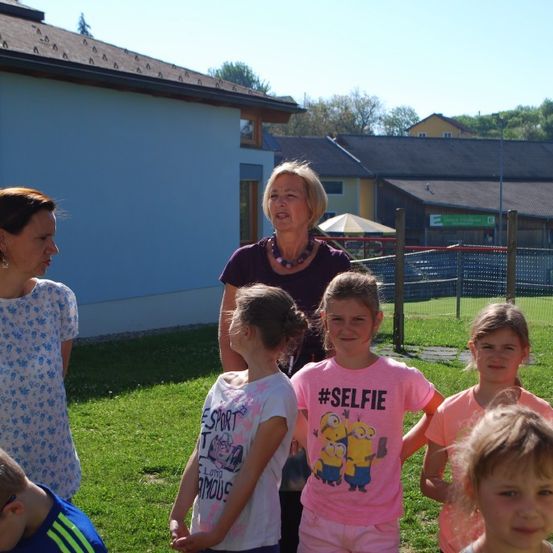Eine Gruppe von Kindern und zwei Frauen stehen auf einem Rasen neben einem Gebäude. Die Frau in der Mitte spricht mit den Kindern. Die Kinder schauen in die Ferne. Dahinter steht ein Zaun mit einem Rasenfeld.