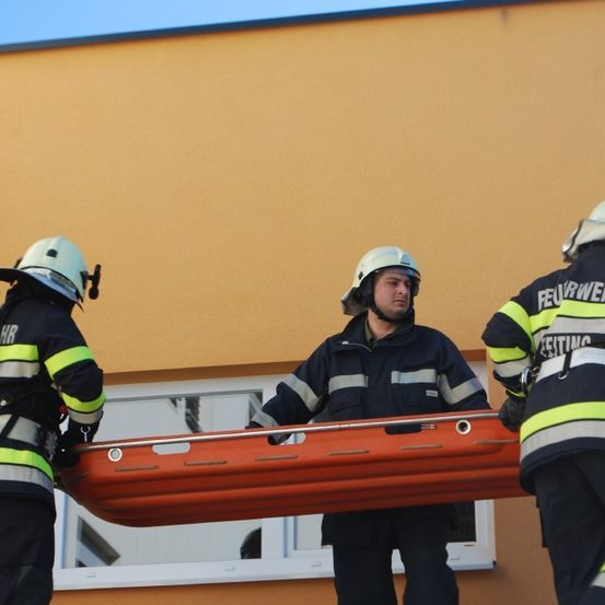 Drei Feuerwehrleute in gelben und schwarzen Uniformen, ausgestattet mit Helmen, tragen eine orangefarbene Trage aus einem Gebäude.
