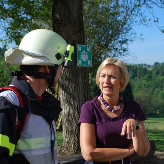 Eine Frau mit blonden Haaren und einer Halskette spricht mit einem Mann, der einen Helm und eine reflektierende Weste trägt, mit einem Baum und einem Schild im Hintergrund.
