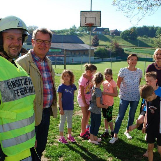 Eine Gruppe von Menschen, einschließlich Kindern und Erwachsenen, versammelt sich auf einem Rasenfeld. Ein Mann in einer reflektierenden Weste sticht hervor, und im Hintergrund befindet sich ein Basketballkorb.