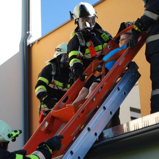 Ein Feuerwehrteam, das Helme und Schutzausrüstung trägt, transportiert vorsichtig eine Person auf einer roten Leiter. Sie stehen auf einem Dach, wobei ein Feuerwehrmann auf der Leiter die Person sicher festhält.