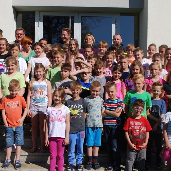 Eine große Gruppe von Kindern und Erwachsenen steht zusammen vor einem Gebäude. Sie lächeln und posieren für ein Foto. Einige tragen Brillen, Turnschuhe und verschiedene Kleidungsdesigns.