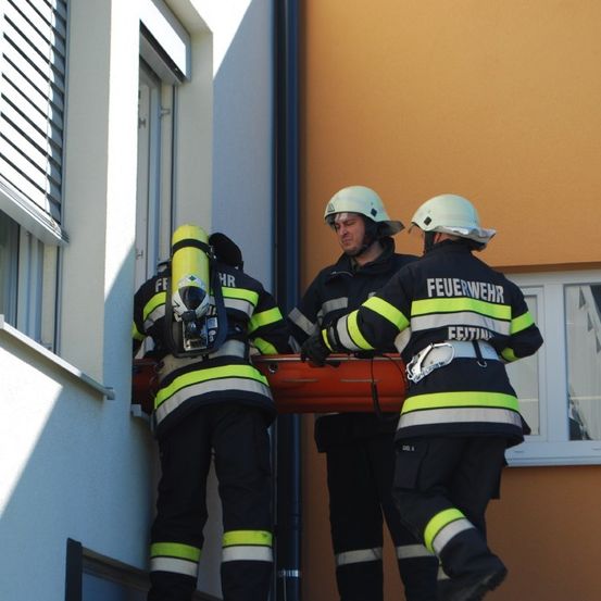 Drei Feuerwehrleute in voller Ausrüstung tragen eine orangefarbene Trage eine Treppe hinunter, wobei einer die Trage hält und die anderen helfen.