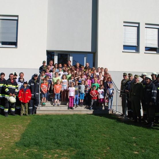 Eine Gruppe Feuerwehrleute, Erwachsene und Kinder posiert für ein Foto vor einem Gebäude.