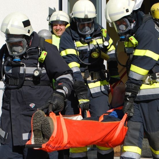 Eine Gruppe von Feuerwehrleuten in Schutzausrüstung trägt eine orangefarbene Trage mit einem sichtbaren Fuß einer Person, sie stehen vor einem Gebäude.