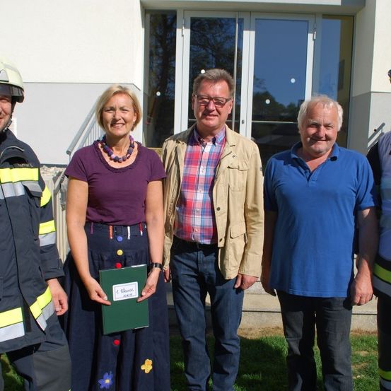 Fünf Personen stehen vor einem Gebäude mit Glastüren. Eine Frau hält eine grüne Mappe. Der Mann rechts trägt ein blaues Poloshirt, und der Mann links trägt eine Feuerwehruniform.