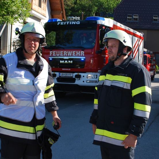 Zwei Feuerwehrleute stehen vor einem roten Feuerwehrauto, einer hält ein Funkgerät, mit Häusern und Bäumen im Hintergrund.
