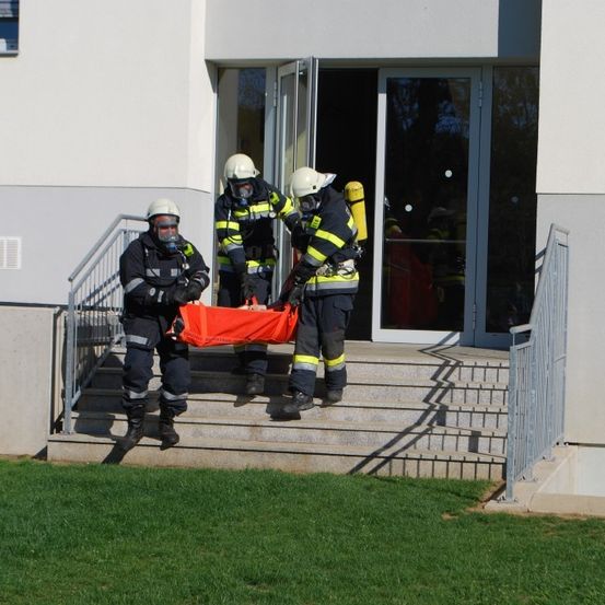 Drei Feuerwehrleute in Schutzausrüstung tragen eine Trage auf den Stufen eines Gebäudes. Sie betreten das Gebäude durch die offene Tür.