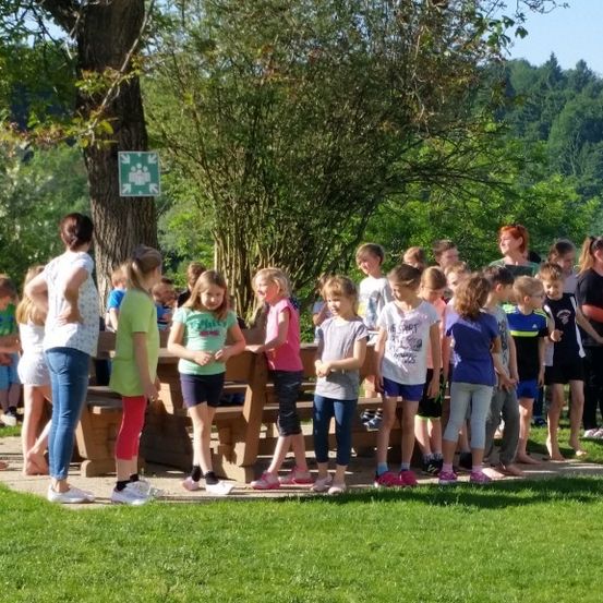 Eine Gruppe von Kindern und Erwachsenen steht in einem Park. Die Kinder stehen vorne und tragen verschiedene farbige T-Shirts. Hinter ihnen befinden sich Holzbänke und ein Baum.