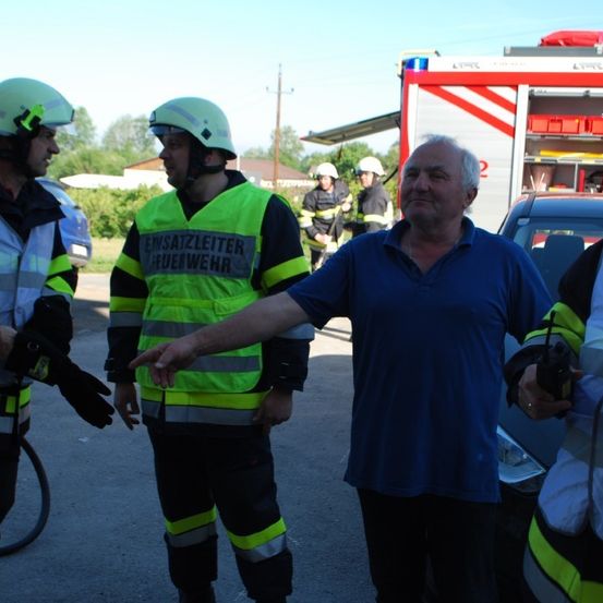 Eine Gruppe von Feuerwehrleuten in voller Ausrüstung unterhält sich mit einem älteren Mann in einem blauen Shirt. Sie sind draußen in der Nähe eines Feuerwehrwagens und eines Fahrrads.