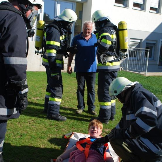 Eine Gruppe von Feuerwehrleuten, die Helme und Uniformen tragen, bereitet einen jungen Jungen auf einer Trage in einem Grasbereich vor. Der Junge lächelt. Dahinter steht ein Mann in der Nähe eines Gebäudes.