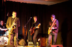 Eine Band spielt auf einer Bühne mit schwarzem Vorhang im Hintergrund. Der führende Saxophonist steht und spielt, während die Gitarristen auf den Knien spielen. Mikrofone und Lautsprecher sind auf der Bühne aufgebaut.