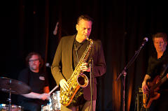 Ein Mann spielt Saxophon auf der Bühne vor einem Mikrofon, während zwei andere Musiker Schlagzeug und Gitarre hinter ihm spielen.