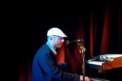 Ein älterer Mann in einem blauen Anzug und einem weißen Hut spielt auf der Bühne ein elektrisches Klavier mit roten Vorhängen im Hintergrund.