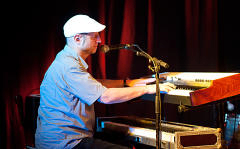 Ein Mann mit einer weißen Mütze spielt ein elektrisches Klavier auf der Bühne, mit einem Mikrofon und einem roten Vorhang im Hintergrund.