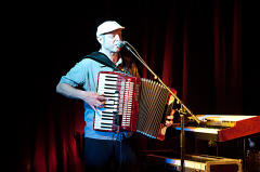 Ein Mann tritt auf der Bühne auf, spielt Akkordeon und singt in ein Mikrofon. Er trägt eine weiße Kappe und ein blaues Hemd. Hinter ihm stehen verschiedene Musikinstrumente, darunter ein Keyboard.