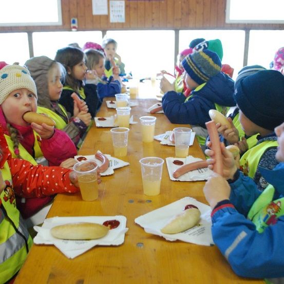 Kinder in Warnwesten essen Hot Dogs an einem langen Tisch. Getränke und Gewürze stehen auf dem Tisch.
