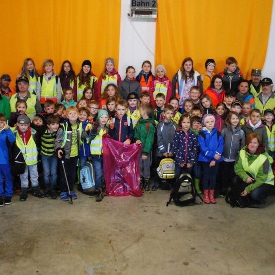 Eine Gruppe von Kindern und Erwachsenen in Sicherheitswesten posiert für ein Foto in einem Raum mit gelben Vorhängen. Einige tragen Mützen, Brillen und Turnschuhe. Eine Person hält eine Plastiktüte.