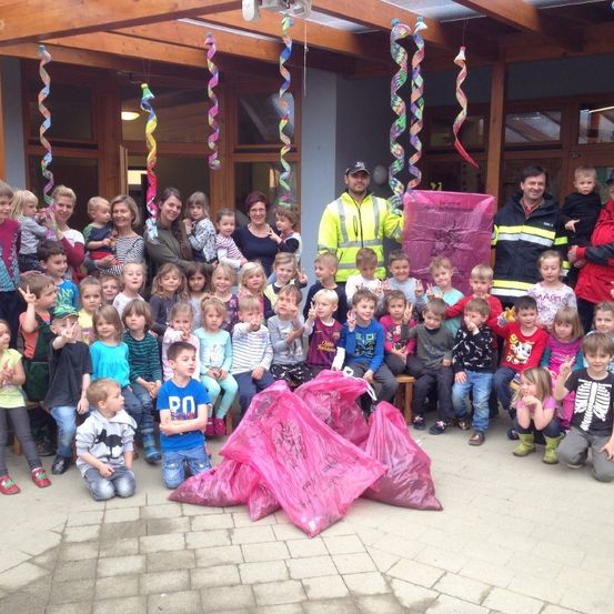 Eine Gruppe von Kindern und Erwachsenen posiert für ein Foto mit zwei Männern, einer hält einen rosa Beutel. Der Ort ist im Freien mit einem Ziegelboden und Dekorationen, die von der Decke hängen.
