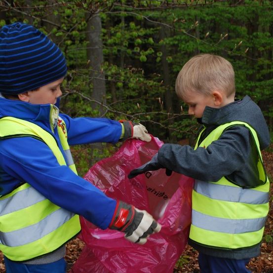 Zwei Jungen sammel im Wald Müll, einer hält einen Beutel, der andere beobachtet. Beide tragen Warnwesten. Bäume mit frischen grünen Blättern umgeben sie.