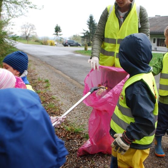 Eine Gruppe von Kindern, die Warnwesten und Handschuhe tragen, sammelt Müll entlang einer Straße, eine Frau in einer gelben Weste hält einen rosa Müllsack.