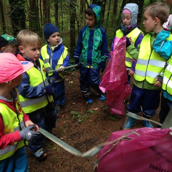 Eine Gruppe von Kindern in Sicherheitswesten im Wald, sammelt Müll mit Stöcken und rosa Tüten.