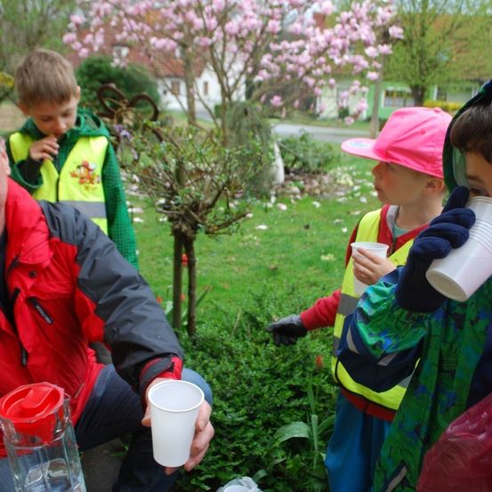 Ein Mann und vier Kinder trinken Wasser aus Tassen im Garten. Sie tragen Warnwesten.