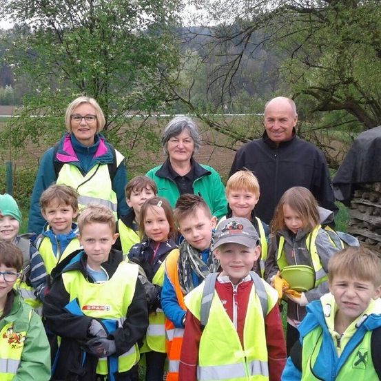 Eine Gruppe von Kindern und zwei Erwachsenen in Warnwesten posiert für ein Foto im Freien. Die Kinder tragen verschiedene Jacken und einige haben Mützen. Hinter ihnen befinden sich Bäume und eine Holzstruktur.