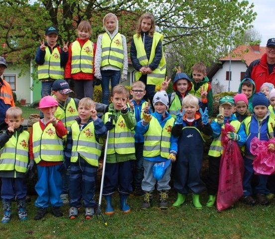 Eine Gruppe von Kindern und Erwachsenen in Warnwesten posiert für ein Foto in einem Park. Einige halten Plastiktüten, und zwei Erwachsene stehen dahinter. Bäume und Häuser sind im Hintergrund.