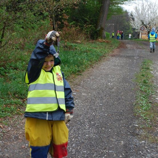 Ein junger Junge in einer Sicherheitsweste und Handschuhen steht auf einem Kiesweg und hält etwas hoch. Dahinter läuft eine Gruppe von Kindern in Sicherheitswesten. Bäume und ein Gebäude sind im Hintergrund zu sehen.