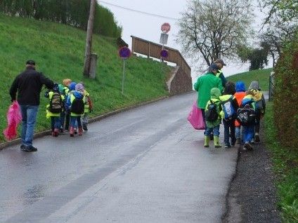Eine Gruppe von Kindern mit Rucksäcken geht an einem nassgewordenen Weg entlang, begleitet von zwei Erwachsenen. Der Weg ist nass, und auf einer Seite befinden sich eine grasbewachsene Anhöhe und Bäume.