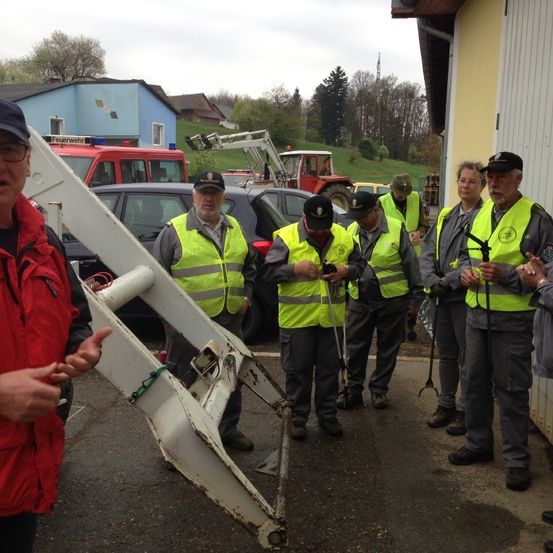 Eine Gruppe von Menschen in gelben Westen und Mützen versammelt sich vor einem gelben Gebäude. Autos und Maschinenfahrzeuge sind hinter ihnen geparkt. Ein Mann in einem roten Jackett spricht zu der Gruppe.