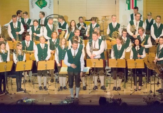 Ein großes Orchester steht auf der Bühne, mit Musikern, die verschiedene Instrumente spielen. Die Orchestermitglieder tragen grüne Westen und Krawatten. Einige stehen, während andere auf Stühlen sitzen. Die Bühne ist mit Musikinstrumenten und Stativen dekoriert.