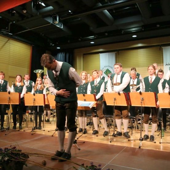 Eine Gruppe von Musikern, gekleidet in grünen Westen, tritt mit verschiedenen Instrumenten auf der Bühne auf. Ein Mann vorne schaut nach unten, möglicherweise singt er.