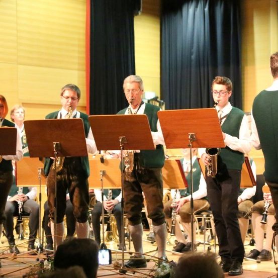 Ein Blasorchester spielt auf der Bühne, mit Musikern, die verschiedene Instrumente wie Saxophone und Trompeten spielen, während ein Publikum von unten zusieht.