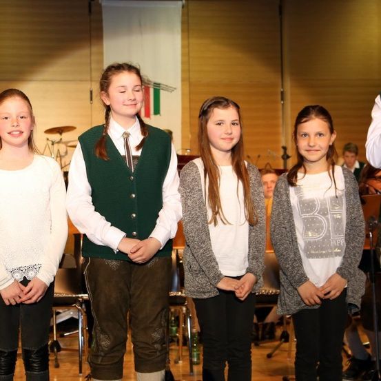 Fünf Mädchen, eine mit einem Mikrofon, stehen zusammen vor einer Bühne, wobei Instrumente und eine Flagge im Hintergrund zu sehen sind.