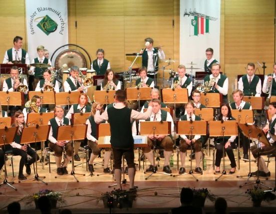 Ein Orchester auf der Bühne mit verschiedenen Instrumenten, darunter Trompeten, Posaunen und Trommeln, tritt vor einer Holzwand mit Bannern auf.