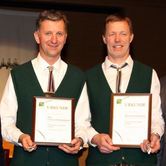 Zwei Männer in grünen Westen und Krawatten halten Urkunden. Der Mann auf der linken Seite lächelt und hält eine Urkunde mit dem Namen Mag Gerhard Monsberger. Der Mann auf der rechten Seite lächelt ebenfalls und hält eine Urkunde mit dem Namen Manfred Ohendrauf.