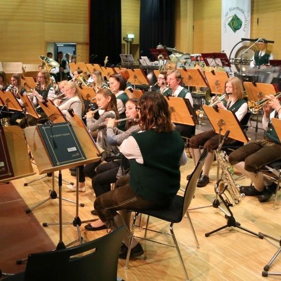 Eine Gruppe von Musikern in grünen Westen und braunen Hosen spielt verschiedene Instrumente in einem Musikraum. Sie sitzen auf Stühlen mit Notenständern vor ihnen.