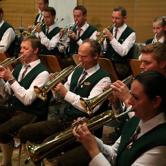 Eine Gruppe von Musikern in grünen Westen und weißen Langarmhemden spielt Trompeten in sitzender Formation. Die Musiker sind in einer Reihe positioniert, mit zwei Frauen an den Enden.
